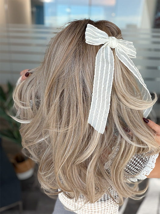 Roxie wearing BROOKLYN by BELLE TRESS in color MILKSHAKE BLONDE R | Mixture of Light sandy brown, highlighted with light ash blonde with a hint of light platinum blonde with medium root color. (styled with bow - not included)